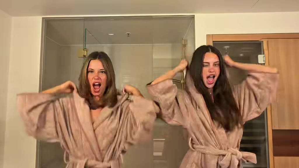 Original frame: two women in tan bathrobes in a bathroom, hands on their hoods