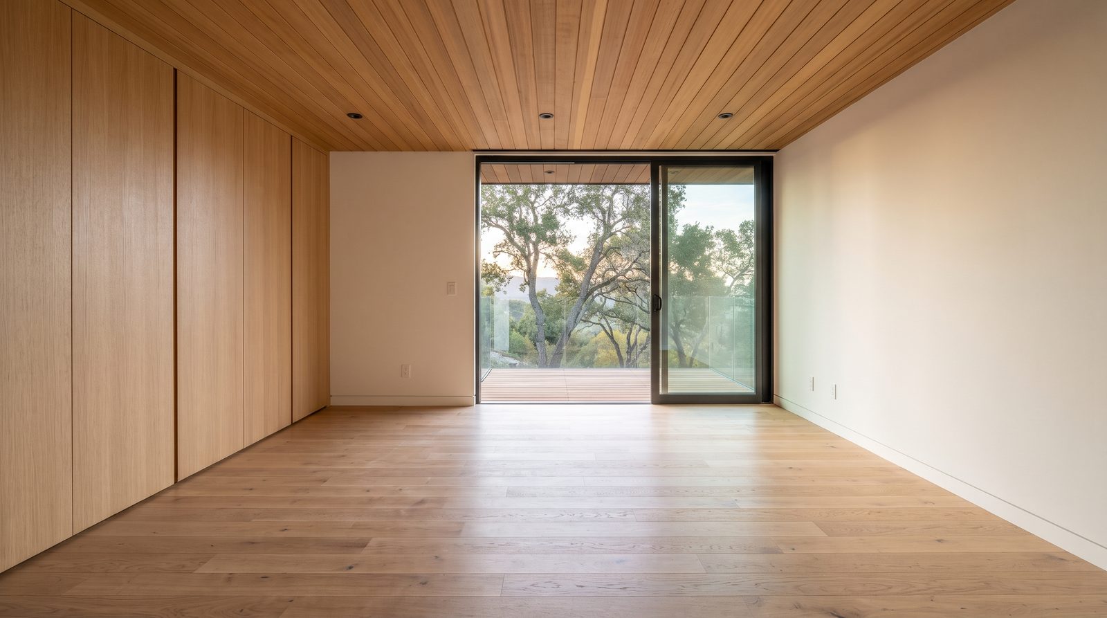 Empty bedroom with oak built-in wardrobes, a slatted wood ceiling and a sliding glass door to a deck with oak trees