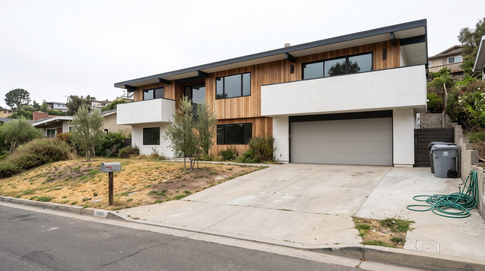 House exterior under a flat white sky with a dry yellow lawn, two trash bins and a garden hose on the driveway