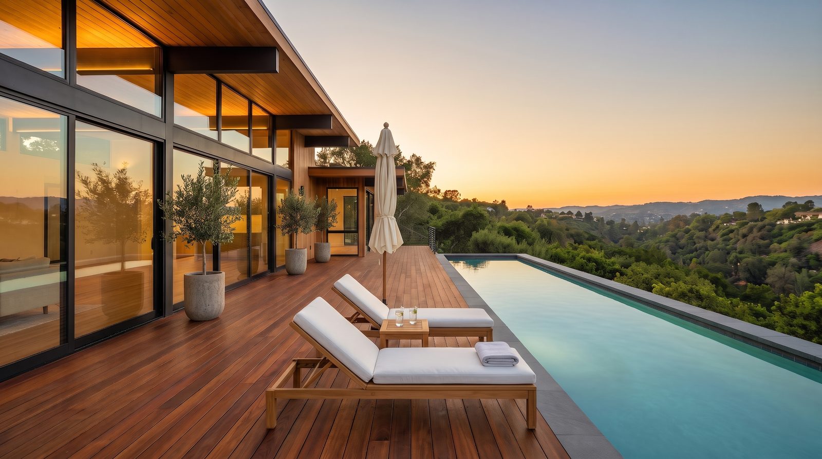 The same pool deck staged with two teak loungers, a side table, a closed umbrella and potted olive trees