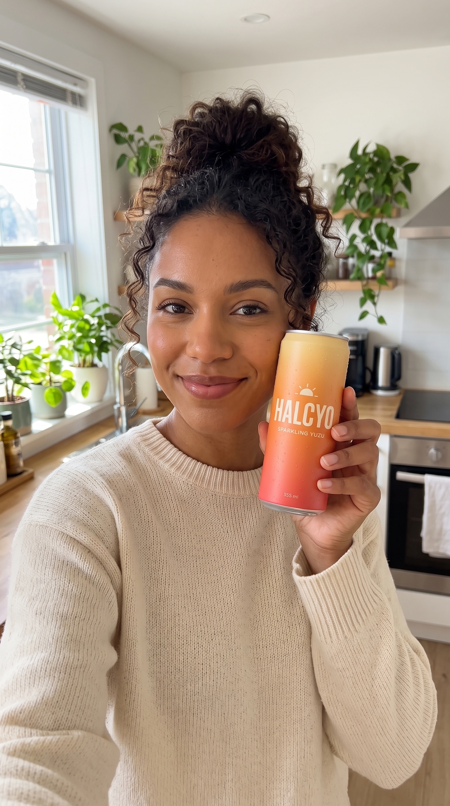 A smiling woman in a kitchen holding a HALCYO can toward a phone camera
