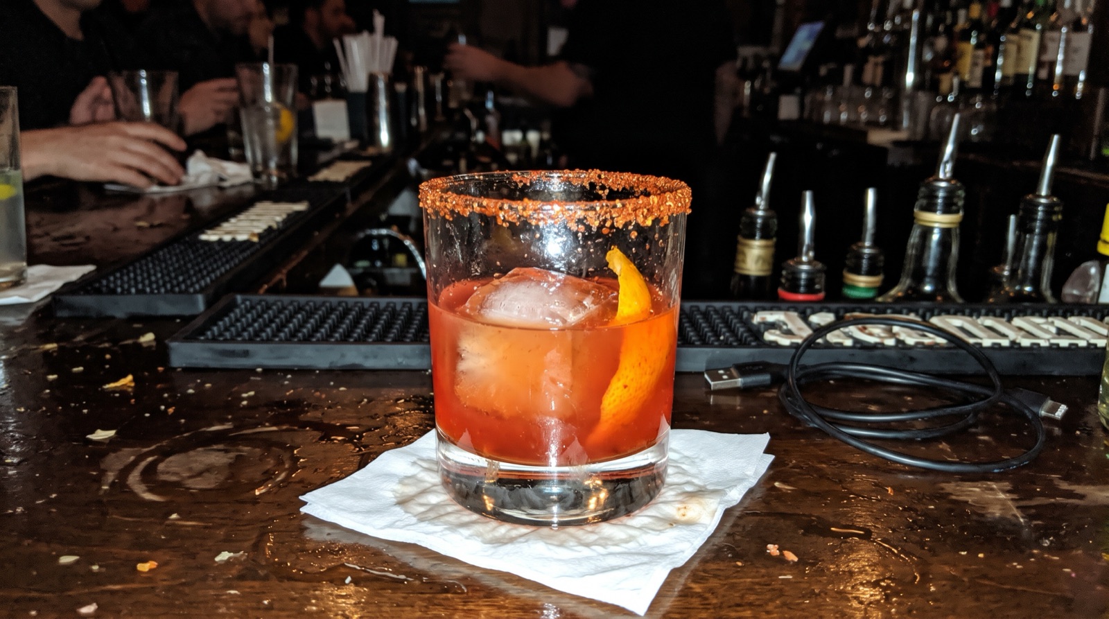 Flash phone photo of a mezcal cocktail on a cluttered bar with a charger cable